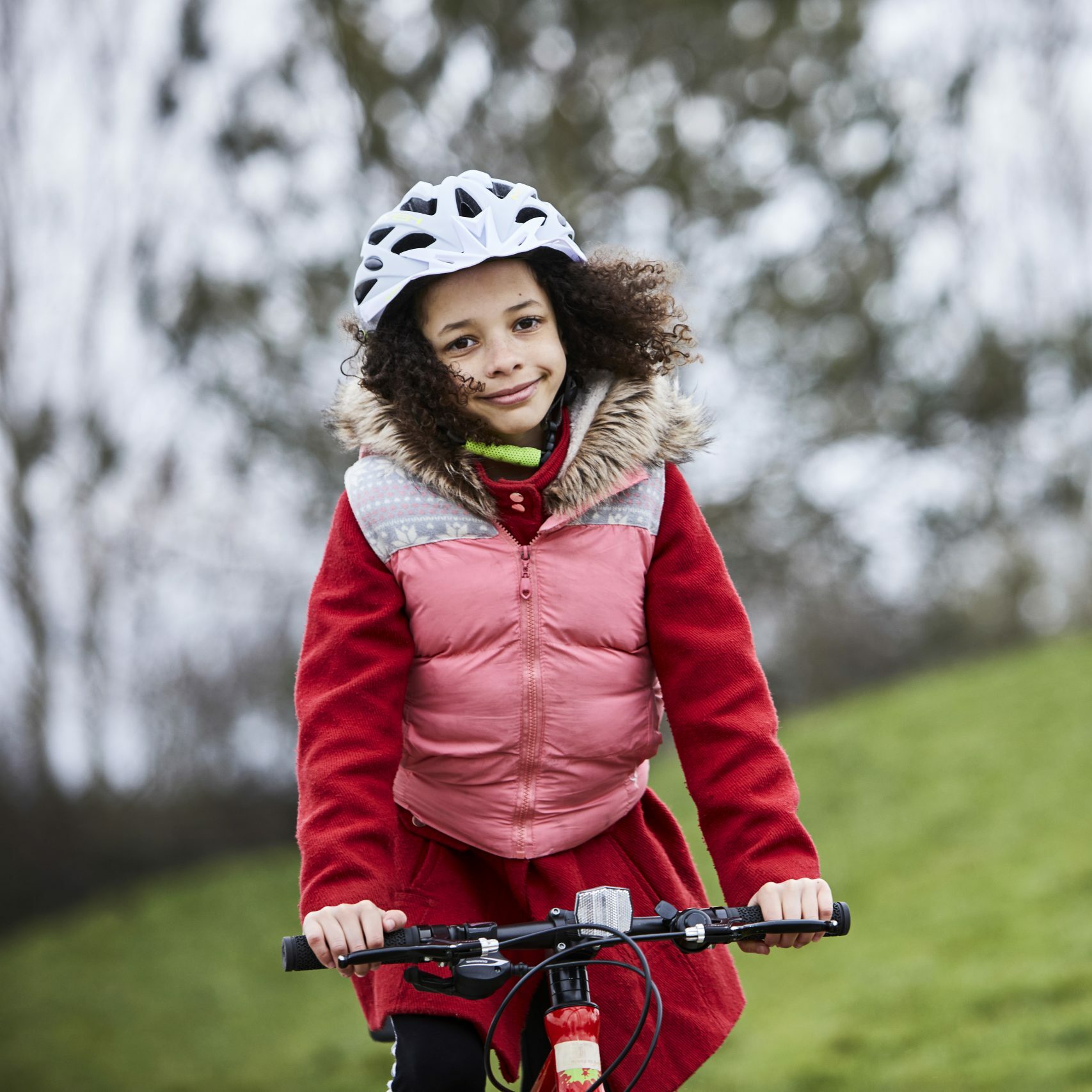 A young girl on her cycle in a park. She has a pink body warmer on over a red coat and a big grin