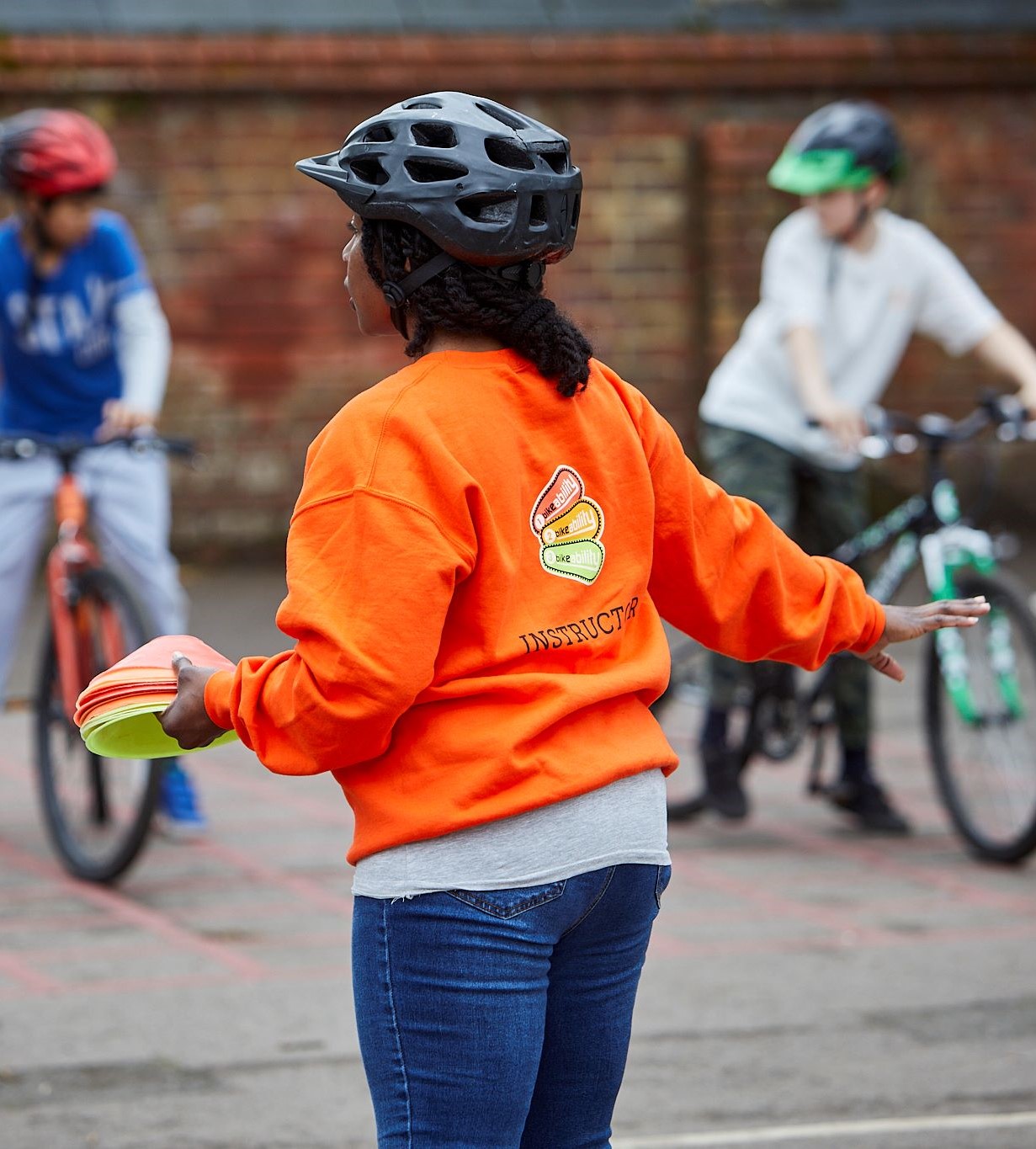 A woman in an orange jumper with instructor on it is talking to some riders in the playground