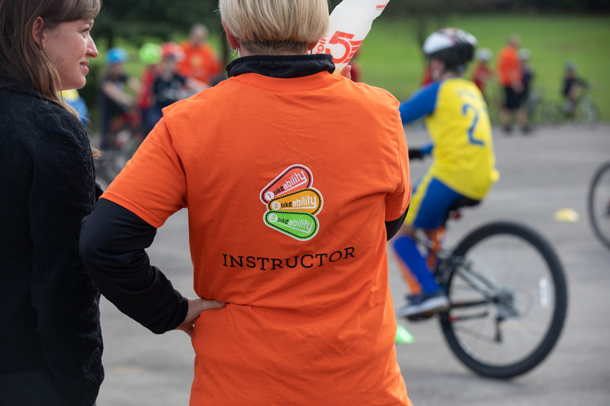 An instructor in the playground watching riders practising. They are wearing a bright orange t shirt with the Bikeability logo and the word Instructor on it