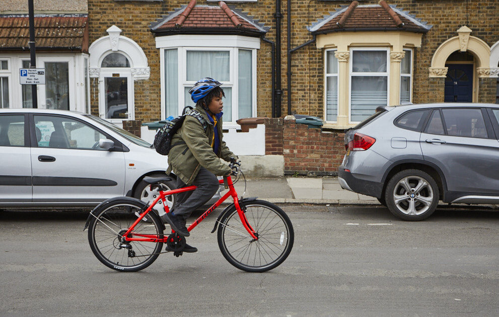 A boy wearing a green jacket and blue helmet cycles along a residential street on a red bike.