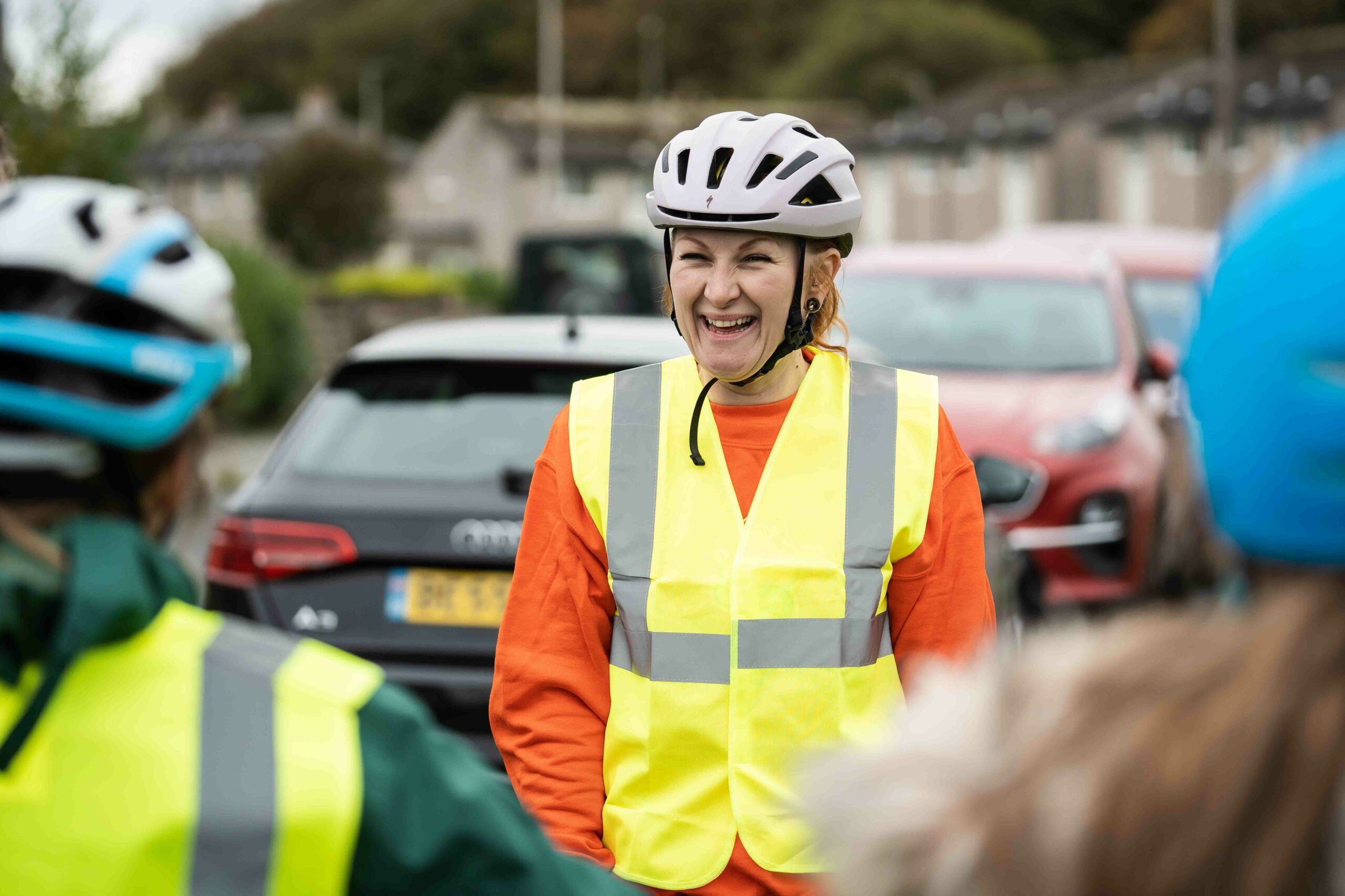 A Bikeability instructor wearing an orange jumper and high vis vest faces the camera smiling. In the foreground are the backs of two children's heads which the camera is looking thorugh.