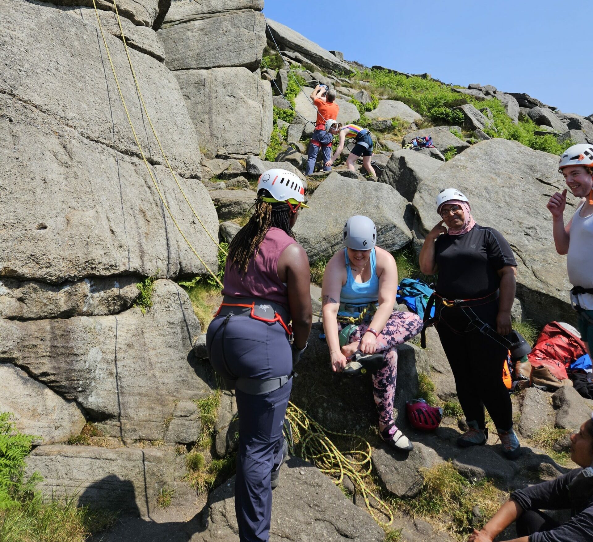 Naida with a group of women preparing to climb a mountain. They are adjusting their shoes while wearing helmets and have climbing ropes attached to them.