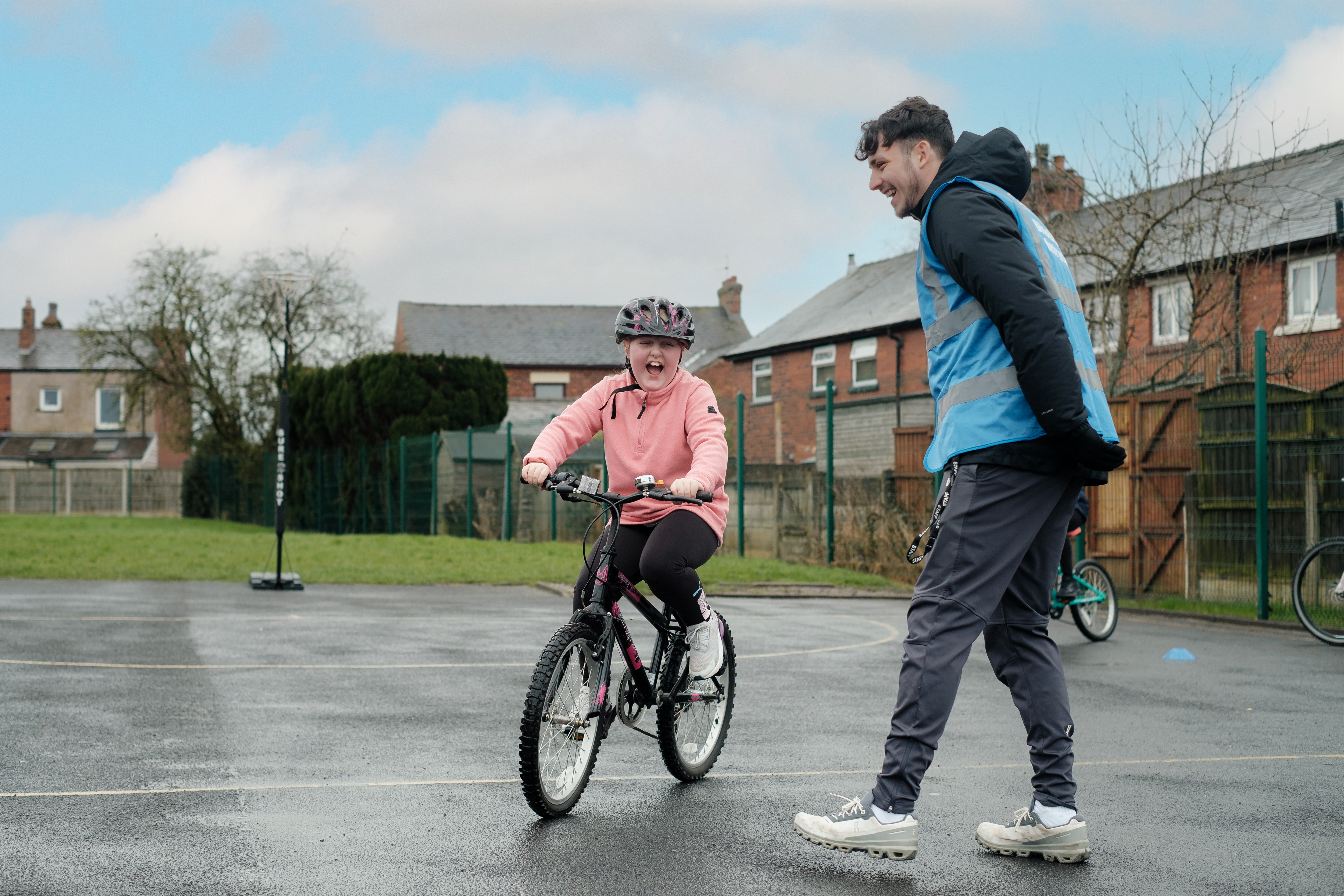 A young trainee is riding her bicycle in a school playground during a Bikeability Level 1 session. Her Bikeability instructor is standing encouragingly beside her. They are both laughing happily and the rider is wearing a safety helmet.