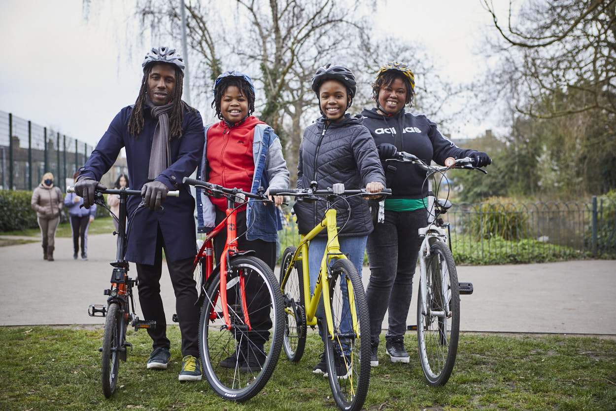 A family consisting of a mum, dad, and two young adolescent children are standing in a park with their bicycles. They are all smiling at the camera and wearing safety helmets.
