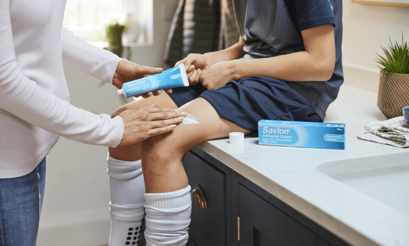 View of a child's knees as they sit on a bathroom counter and an adult, holding a tube of savlon, rubs the medication into a small scrape