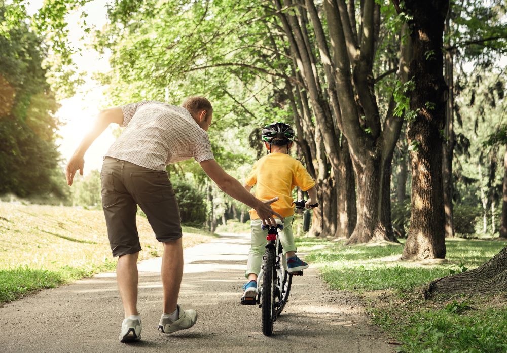 A young child is pushed on his cycle by an adult in the park