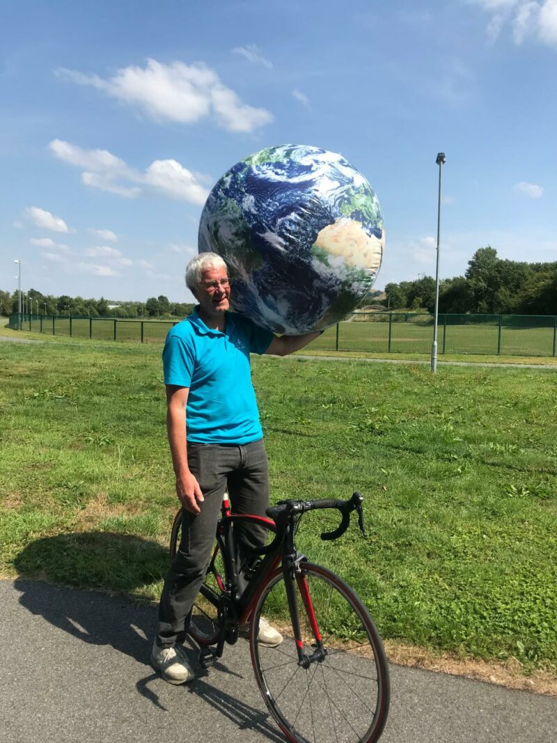 Instructor Adrian is standing with his bike. He is carrying a large model of the earth on his shoulder.