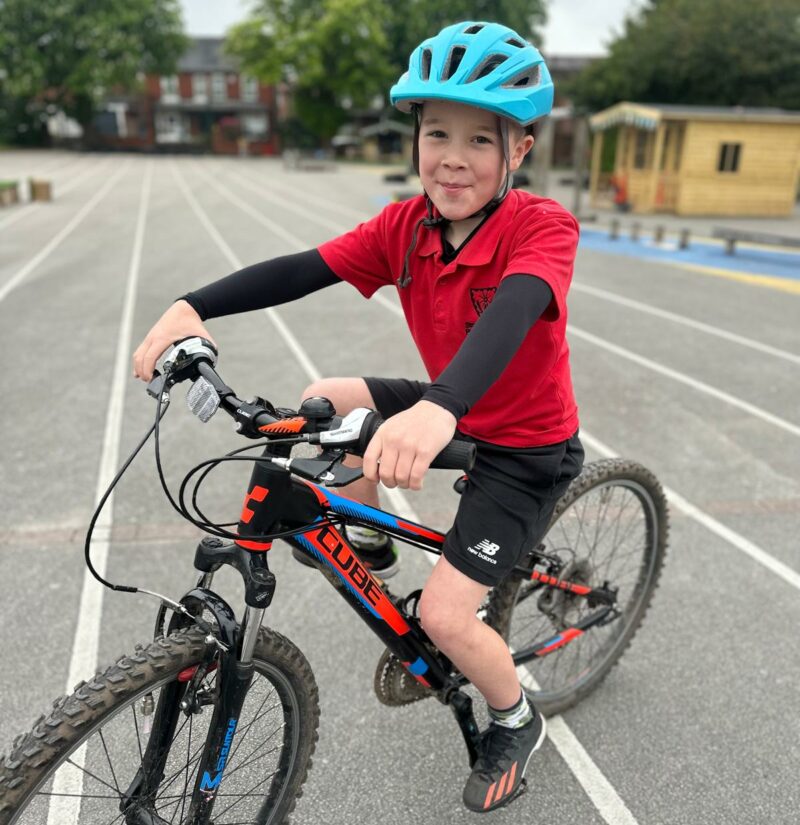 Young cyclist of the Year 2024 Harvey on his bike. He is wearing a blue helmet and a red t shirt and smiling