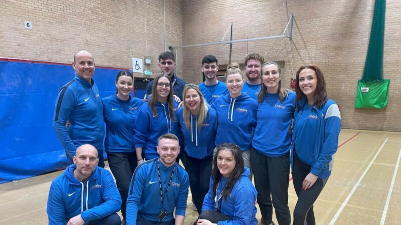 The team from Chorley SSP. There are 13 people, all wearing blue tops and standing in a gym together smiling for the camera