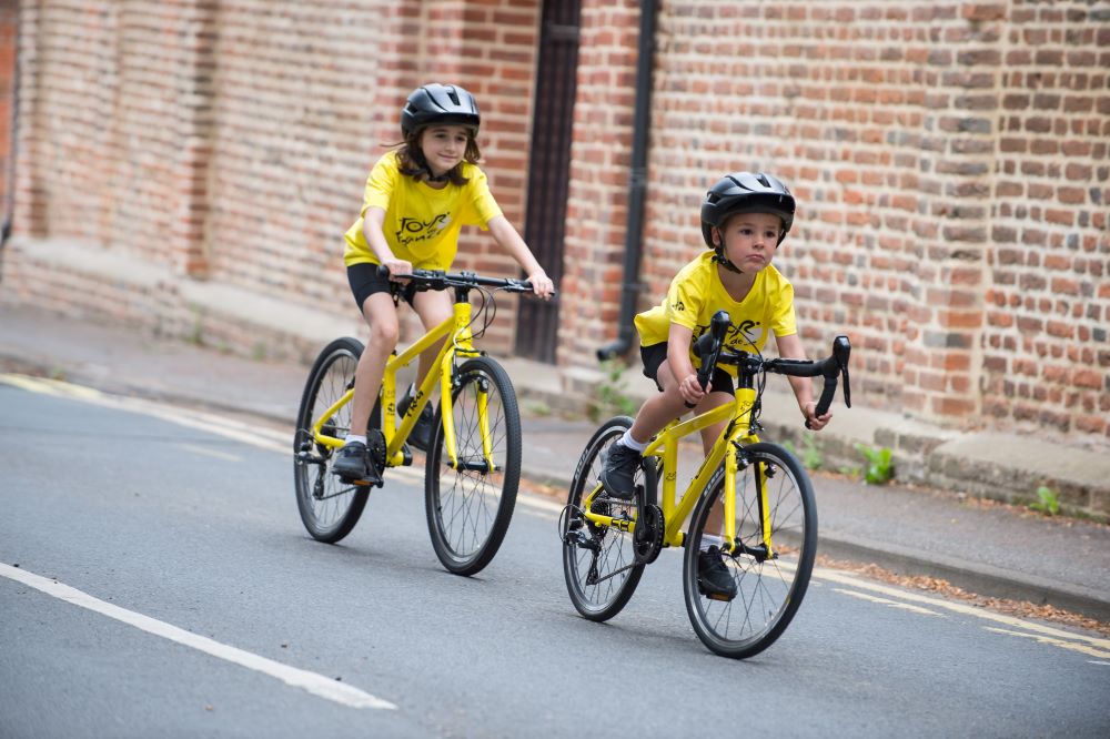 Two children, a boy and a girl, cycling on Frog Tour de France branded bicycles. They are both wearing yellow Tour de France branded t-shirts and cycling along a suburban road.
