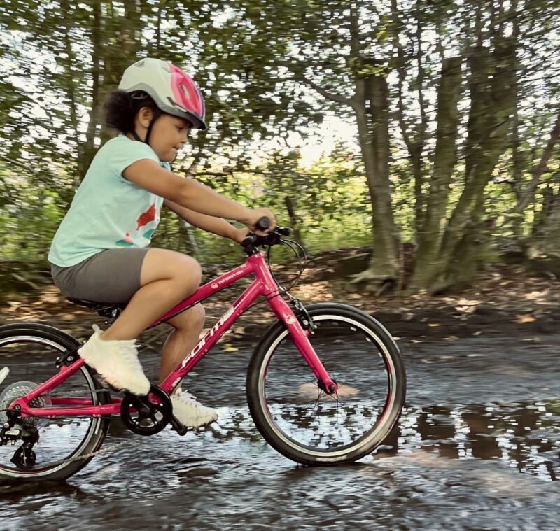 A girl on a red bicycle in a green t shirt is riding her bike through a big puddle