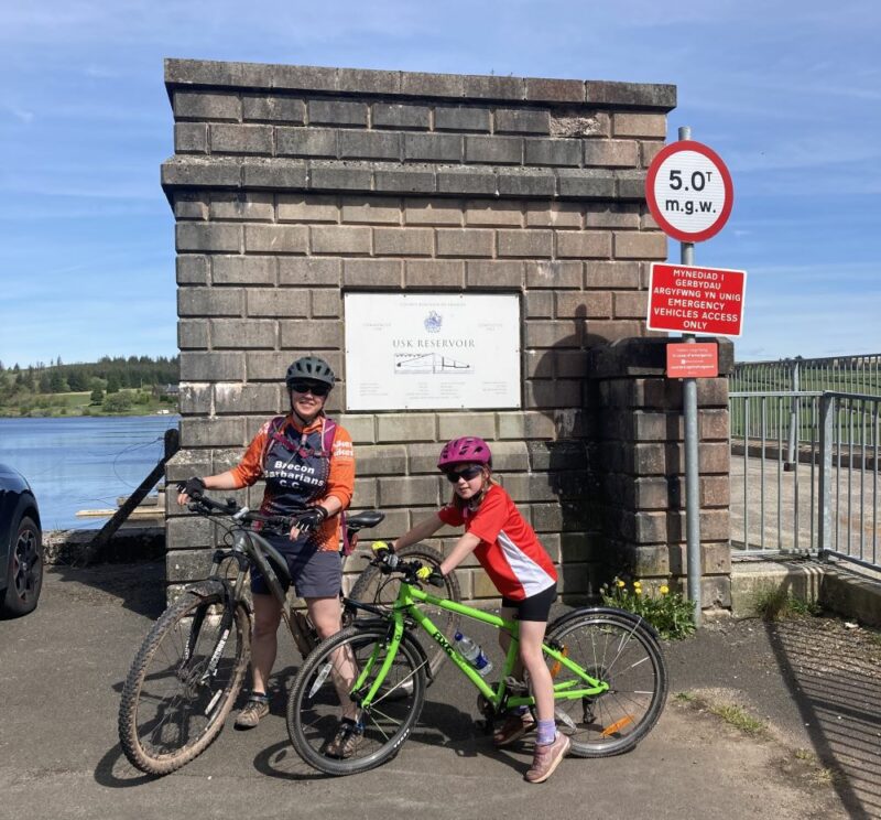 A young girl and her mum are on their cycles in front of a brick structure that says Usk Reservoir on it