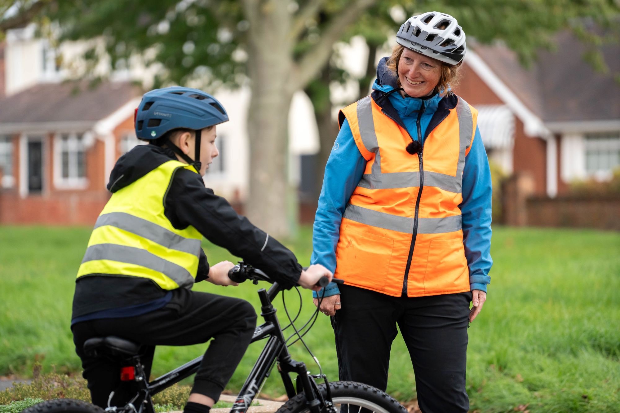 A Bikeability instructor in an orange hi-vis vest smiles as she watched a rider on a cycle in a yellow hi-vis vest and helmet practising