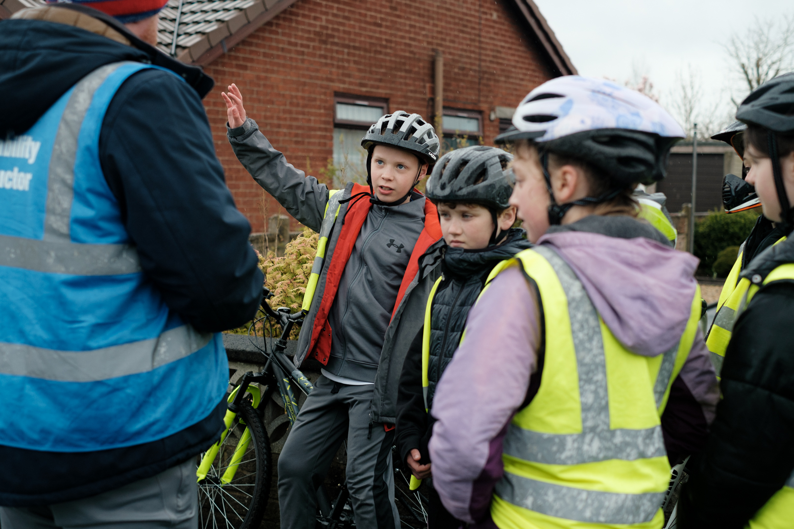 A Bikeability rider raising his hand as an instructor ask him a question. They are in the playground and surrounded by other Bikeability riders taking part in the lesson.