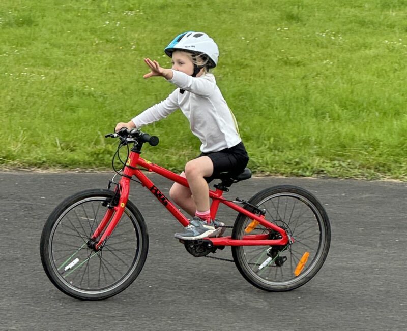 A young boy on a red bicycle is riding and signalling with his left hand to show his is planning to turn left