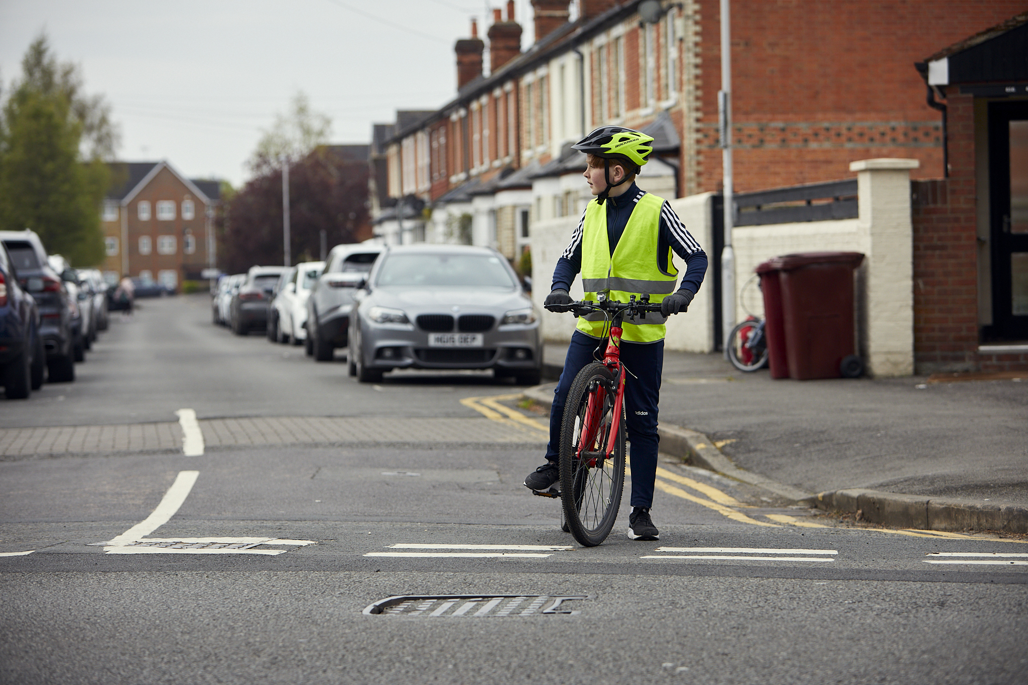 A young boy on his cycle at a junction. He is stopped and looking to his right to check for traffic before moving. He is riding a red cycle and wearing a safety helmet and a hi-vis vest.