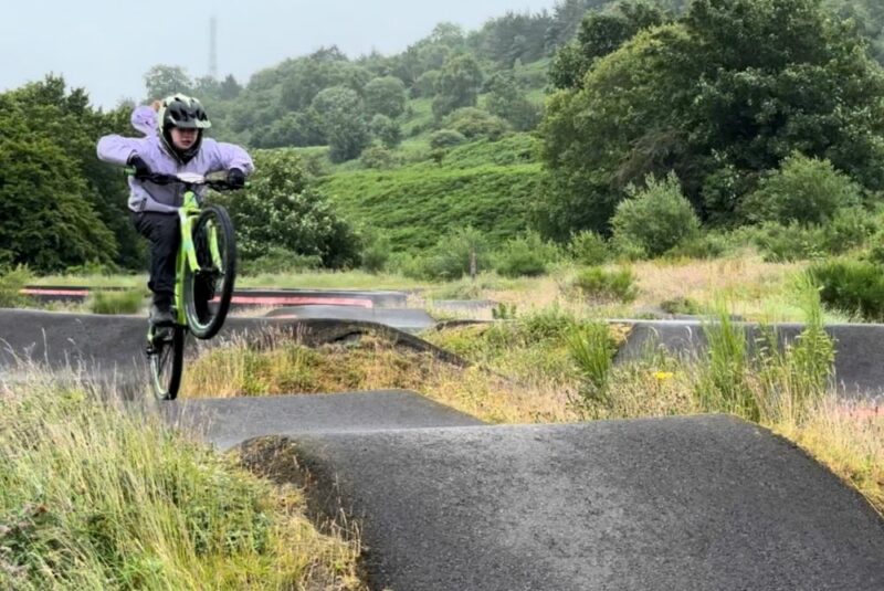 A girl on a green cycle is jumping up a hump on a pump track