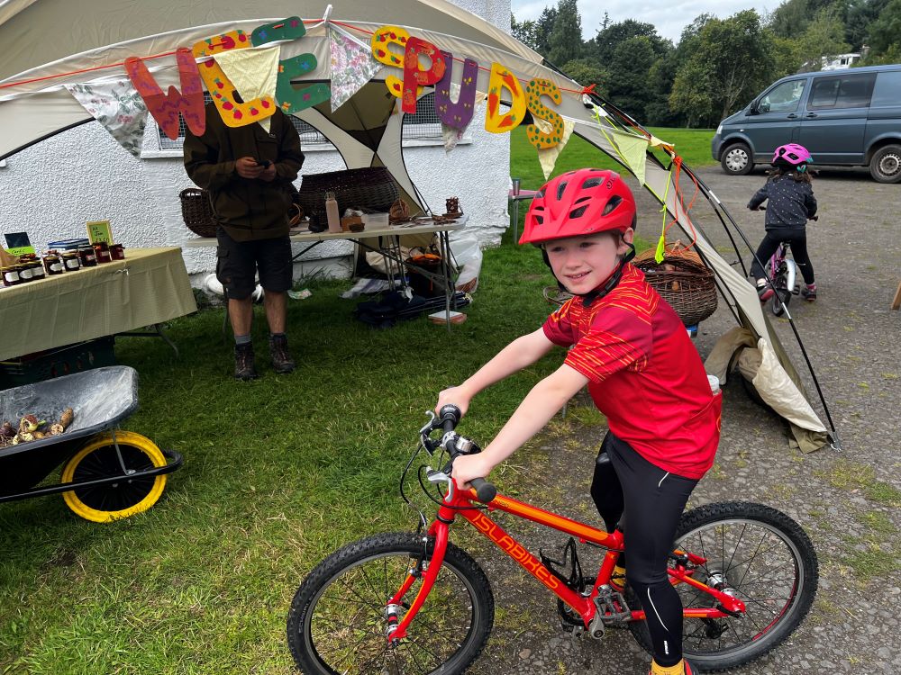 A boy in a red t shirt is on a red cycle in front of a marquee with bunting decorating it