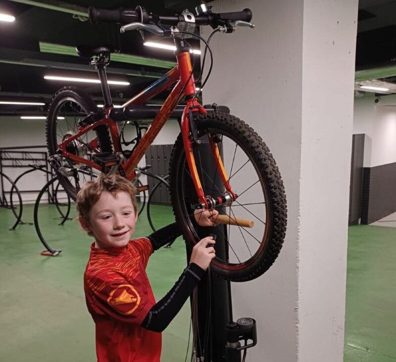 A boy is checking his wheel on his cycle which is on a stand. He is wearing a red top and smiling