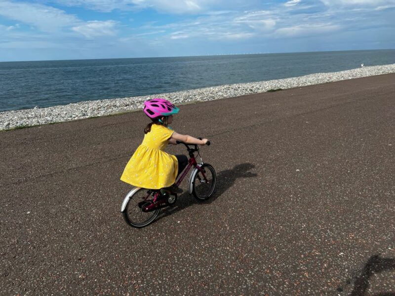 A young girl in a yellow dress is cycling along a beach with the sea in the background