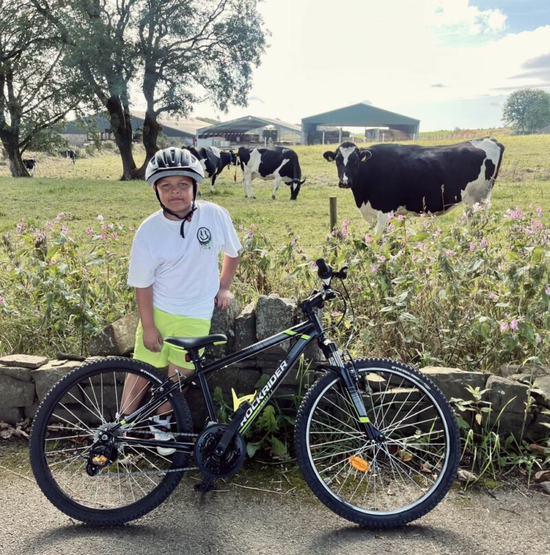 A young boy with his cycle standing in front of a field of cows/ He has a big grin on his face