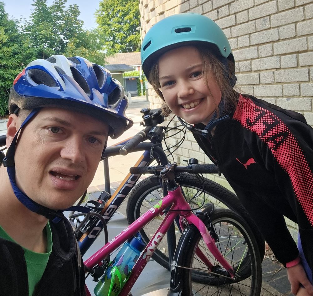 A girl and her dad and smiling at the camera. They are wearing safety helmets and their bikes are in the background