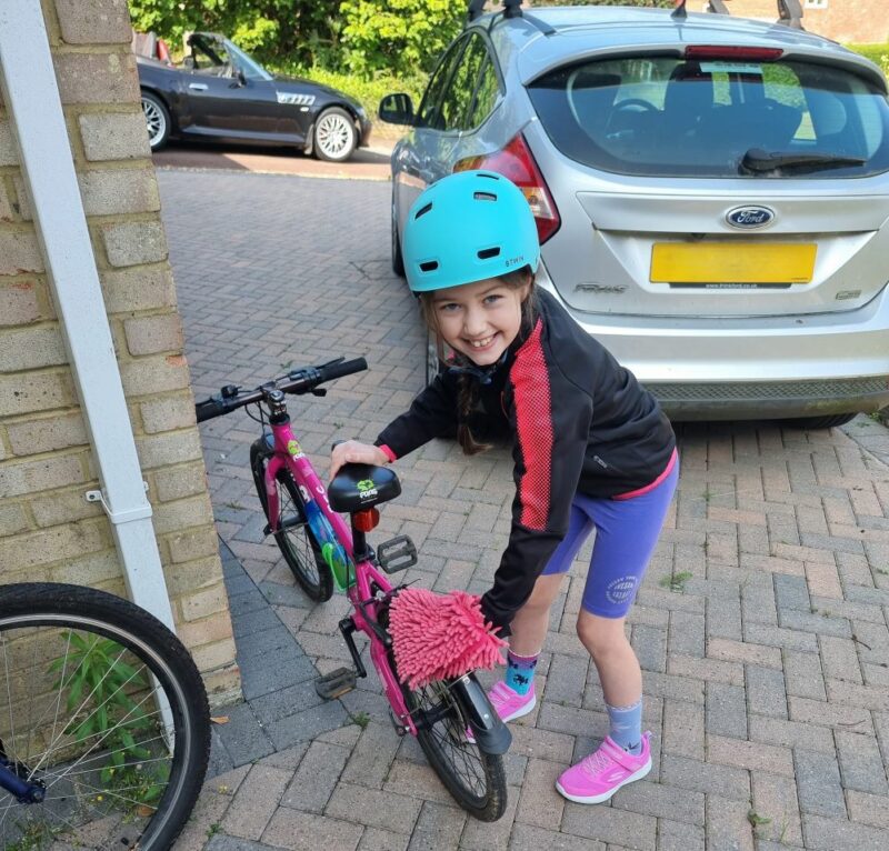 A girl is cleaning her bicycle. She is wearing a blue helmet and looking at the camera and smiling