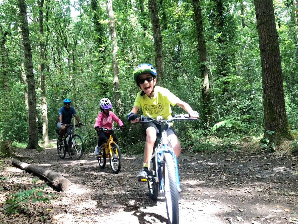 A family are cycling through some woods. A boy i at the front is smiling and laughing as he cycles towards the camera