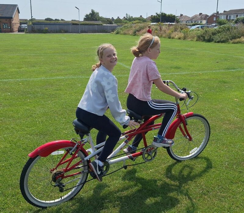 Two girls are riding a bright red tandem bicycle. The girl on the back is looking at the camera and smiling