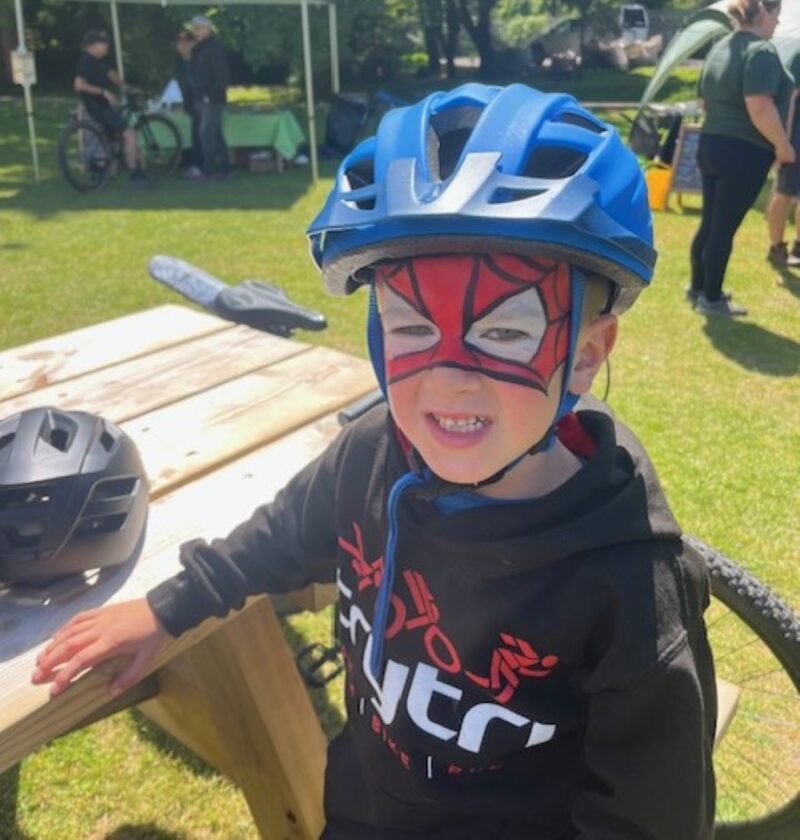 A young boy wearing a cycle helmet is sitting at a bench smiling. He has Spiderman face paint on