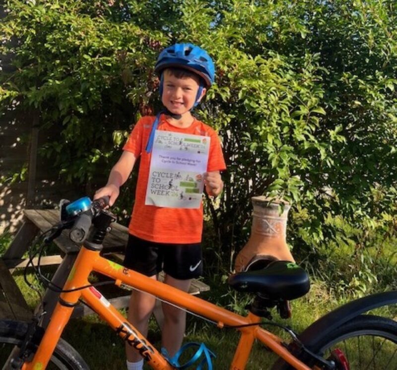 A boy in an orange t shirt with a matching orange bicycle is holding up a print out of his Cycle to School Week pledge