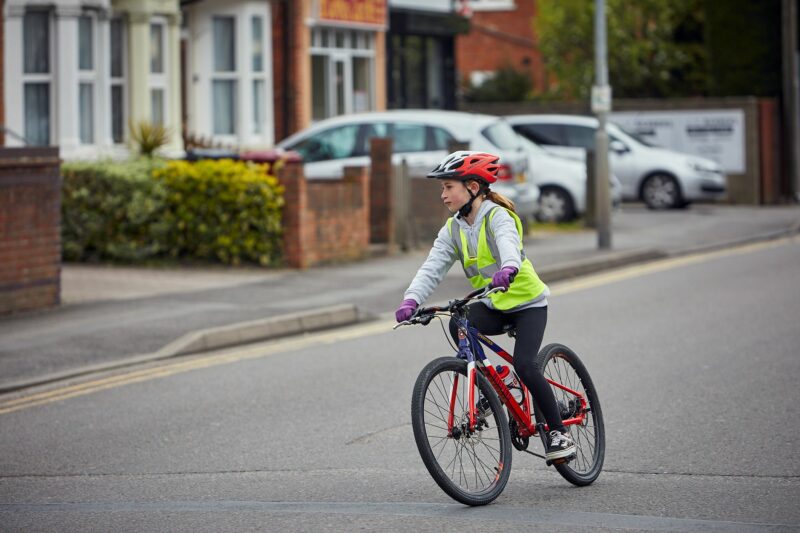 A young girl cycling along the road wearing a helmet and a hi vis vest