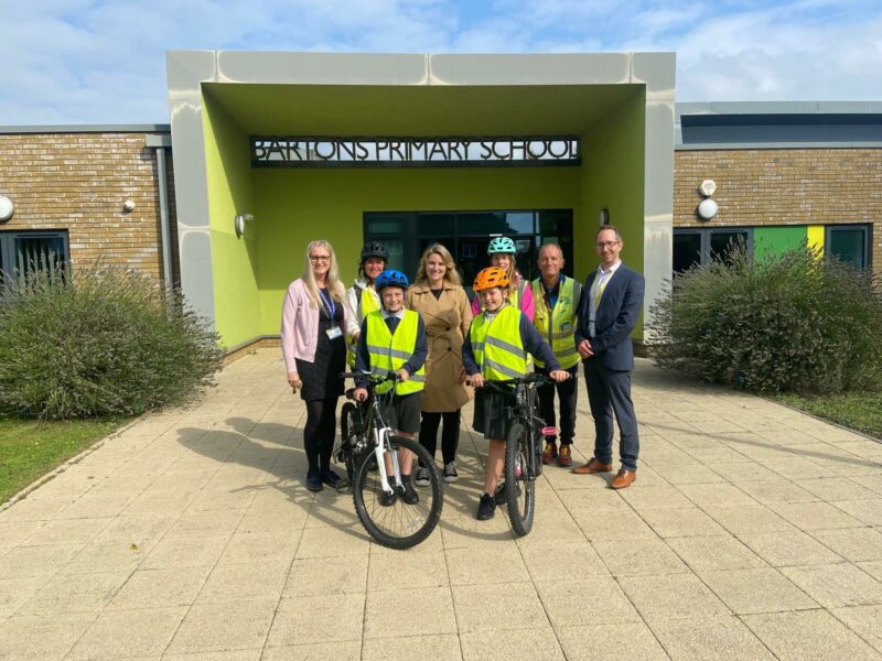 A group of teachers and students standing in front of the entrance to Bartons Primary School. The two students at the front have bikes with them.