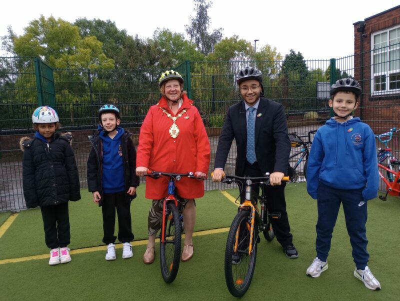 Lord Mayor of Stoke-on-Trent Lyn Sharpe with a teacher and students in a playground. They are all wearing helmets and the Mayor and teacher are on bikes