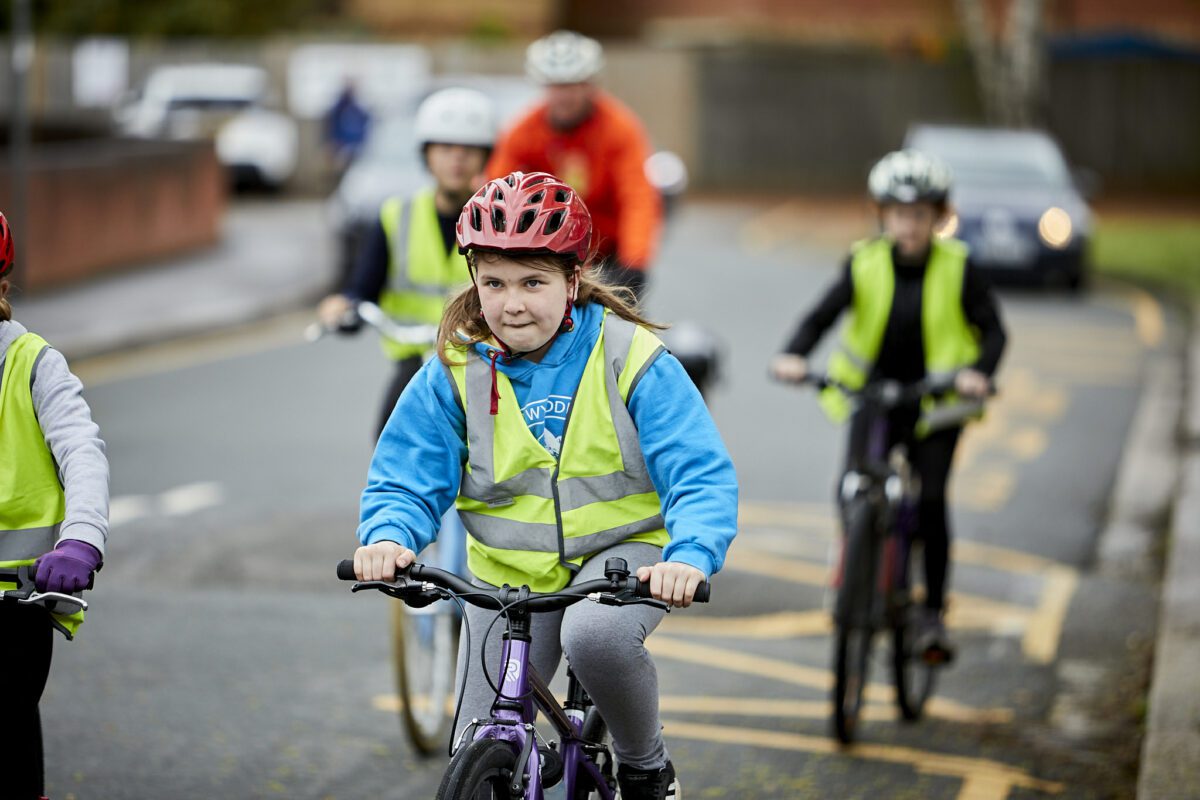 A child is cycling on a road with fellow Bikeability trainees. An instructor can be seen following in the background.